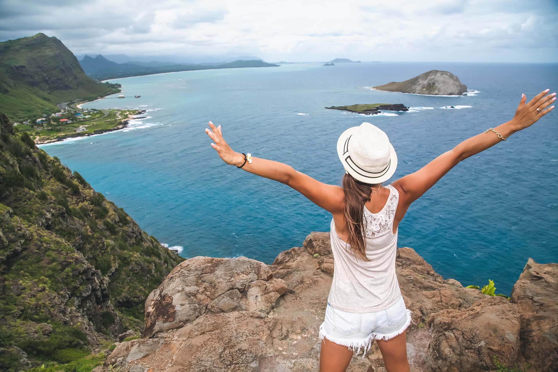 A young woman with her arms stretched out standing on a cliff overlooking the Hawaiian coastline